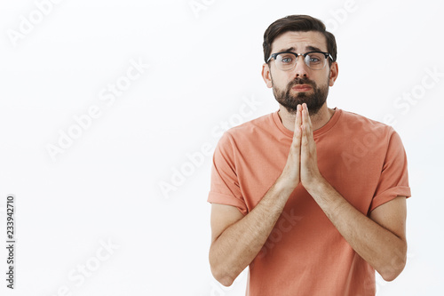 Please I need you help. Silly begging insecure nerdy guy in glasses stooping holding hands in pray gazing with cute sad expression at camera asking favor or lending money posing against gray wall