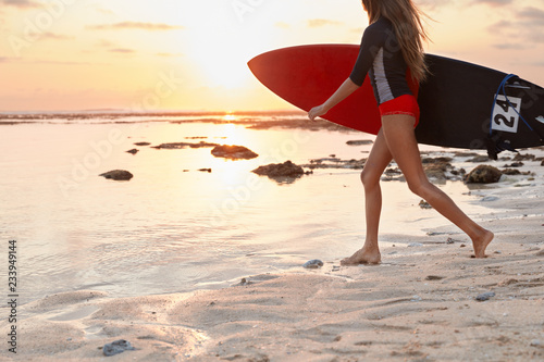 Canvas Print Cropped image of active woman dressed in swimsuit, carries surf board, walks on coastline against sunshine, ready to hit waves, travels in paradise place
