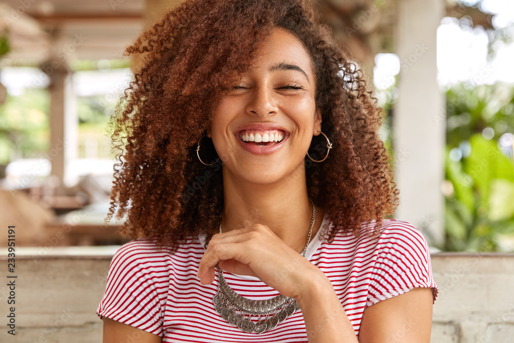 Headshot of glad woman laughs and smiles broadly, expresses positive emotions, has hilarious expression, wears round big earrings and necklace, spends time in cafe. Afro American teenager indoor - obrazy, fototapety, plakaty 
