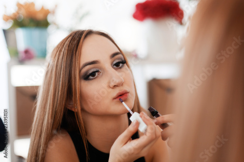 Portrait of a young girl who doing a makeup applying liquid lipstick to her lips looking in the mirror. Shallow focus and blurred background.