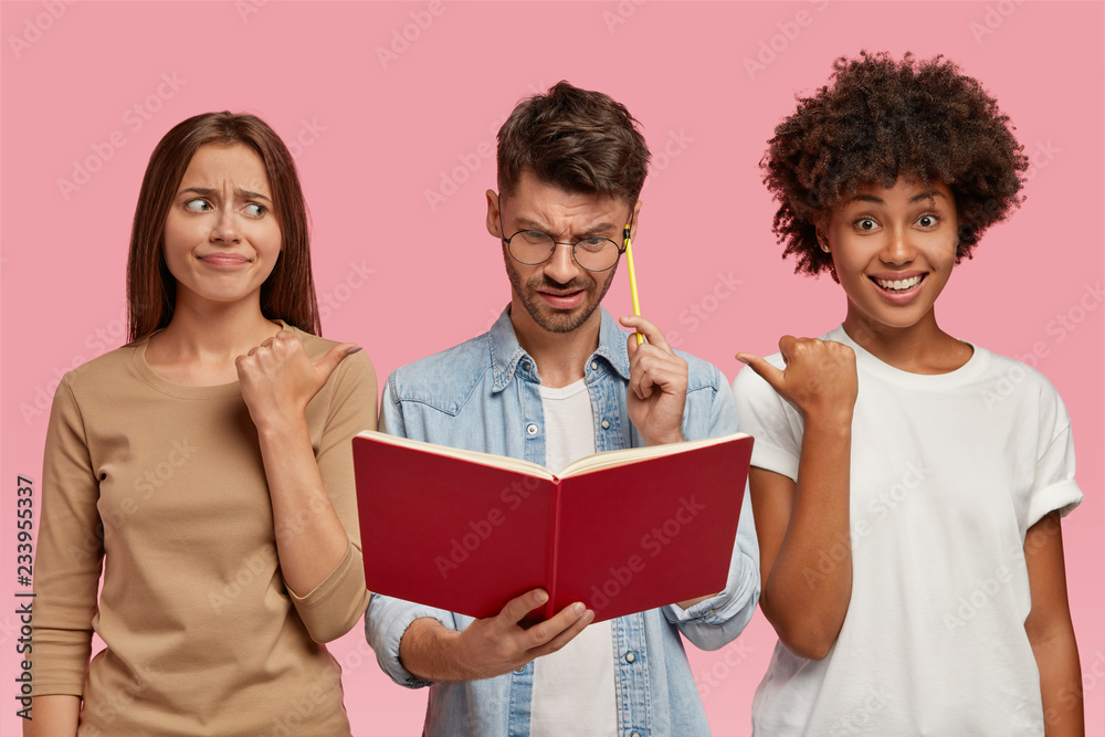 Indoor shot of Caucasian and African women point at their nerd friend who looks in book with puzzlement, learns information, being busy with studying, isolated over pink background. Three students Indoor shot of Caucasian and African women point at their nerd friend who looks in book with puzzlement, learns information, being busy with studying, isolated over pink background. Three students