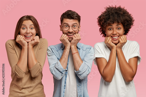 Photo of three multiethnic friends hold chins, watch comedy together during leisure time, wear casual clothes, stand next to each other over pink background. Family portrait. Two sisters and brother