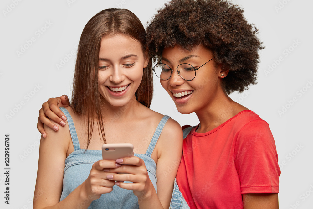 Photo of pretty two multiethnic sisters make booking together on web page via cell phone, watch video online, embrace, isolated over white background. Afro girl has fun with best friend indoor Photo of pretty two multiethnic sisters make booking together on web page via cell phone, watch video online, embrace, isolated over white background. Afro girl has fun with best friend indoor