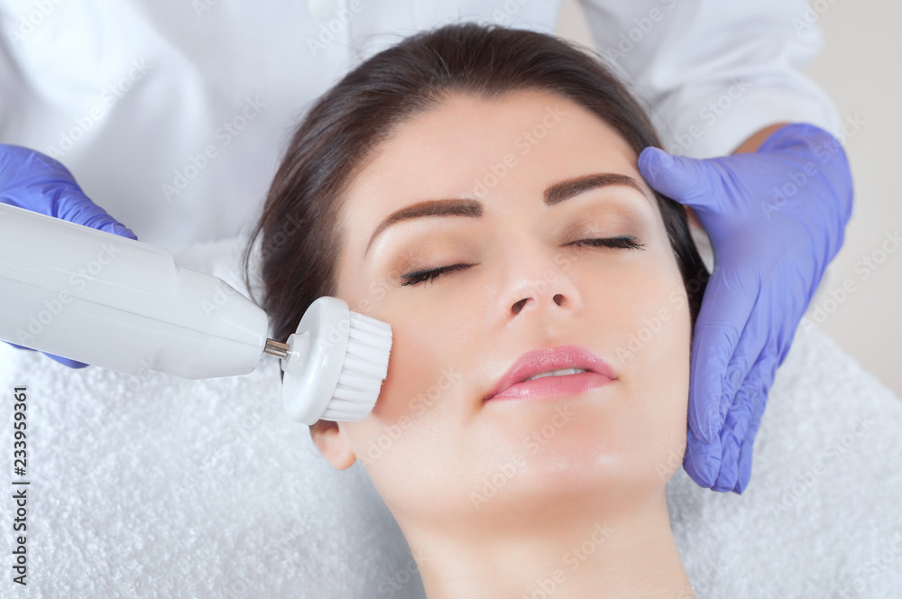 The cosmetologist makes the Hardware face cleaning  procedure with a soft rotating brush of a beautiful, young woman in a beauty salon. Cosmetology and professional skin care.