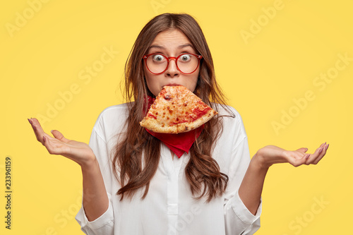 Foto Surprised Caucasian woman keeps slice of pizza in mouth, spreads hands with hesi