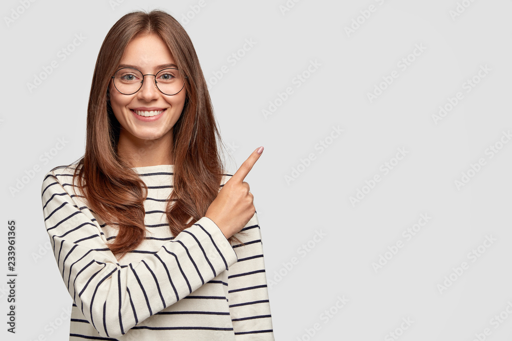 Waist up shot of pretty Caucasian woman with cheerful expression, points with index finger at blank copy space, dressed in striped sweater, shows free space at upper right corner for your promotion - obrazy, fototapety, plakaty 