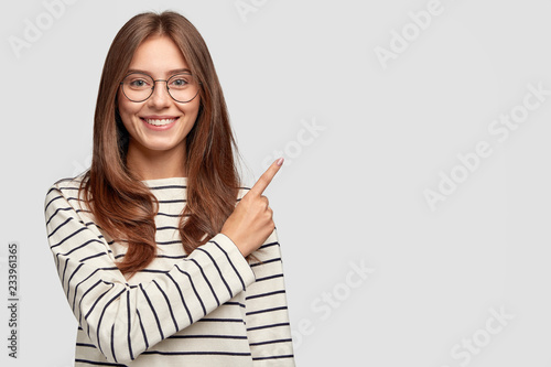 Waist up shot of pretty Caucasian woman with cheerful expression, points with index finger at blank copy space, dressed in striped sweater, shows free space at upper right corner for your promotion photo