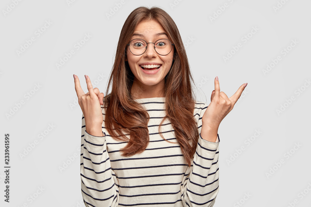 Satisfied woman with toothy smile, raises both hands and shows rock n roll sign, dressed in striped jumper, models against white background, listens heavy music, likes rock. Hipster enjoys music Satisfied woman with toothy smile, raises both hands and shows rock n roll sign, dressed in striped jumper, models against white background, listens heavy music, likes rock. Hipster enjoys music