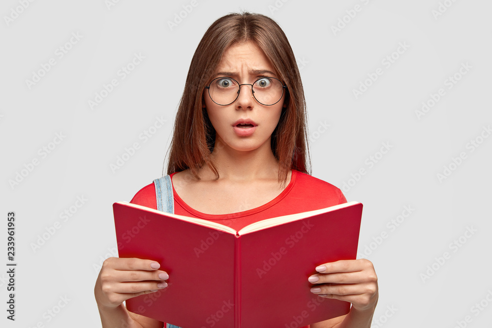 Photo of stupefied emotional young Caucasian woman looks with stupefaction at camera, holds red book, has to learn much for next lesson, wears round spectacles, isolated over white background Photo of stupefied emotional young Caucasian woman looks with stupefaction at camera, holds red book, has to learn much for next lesson, wears round spectacles, isolated over white background