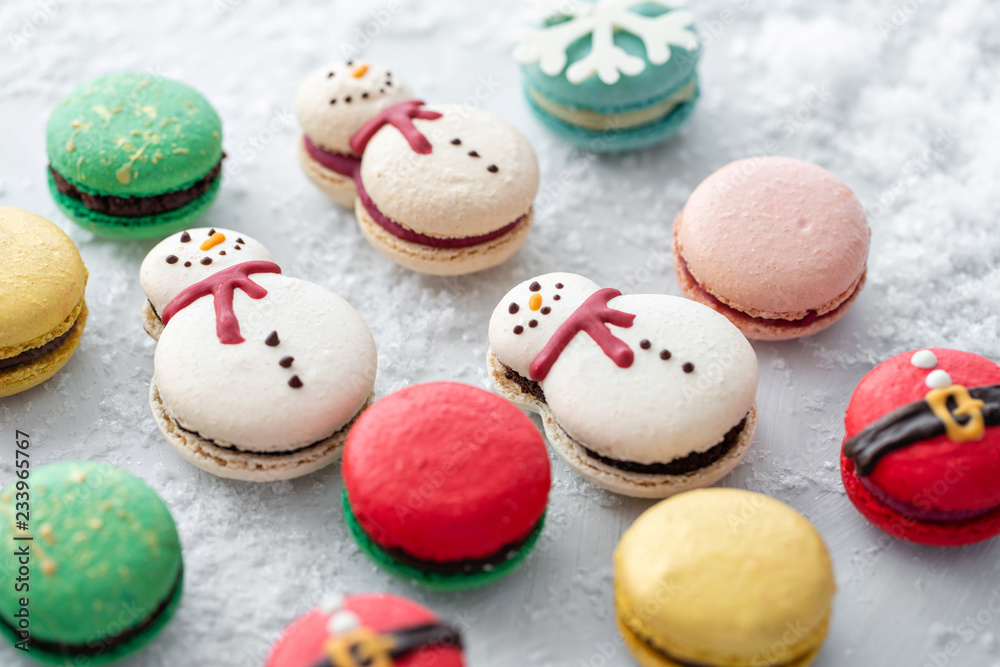 Fototapeta premium Variety of sweet macaroons on gray table sprinkled with snow. Modern european French cuisine. Christmas theme, Merry Christmas card. New year mood. lights of garland bulbs