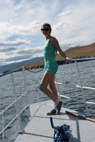 In the summer, on a boat on the water surface, the girl stands aft in Shert and mate against the backdrop of mountains, sky and water