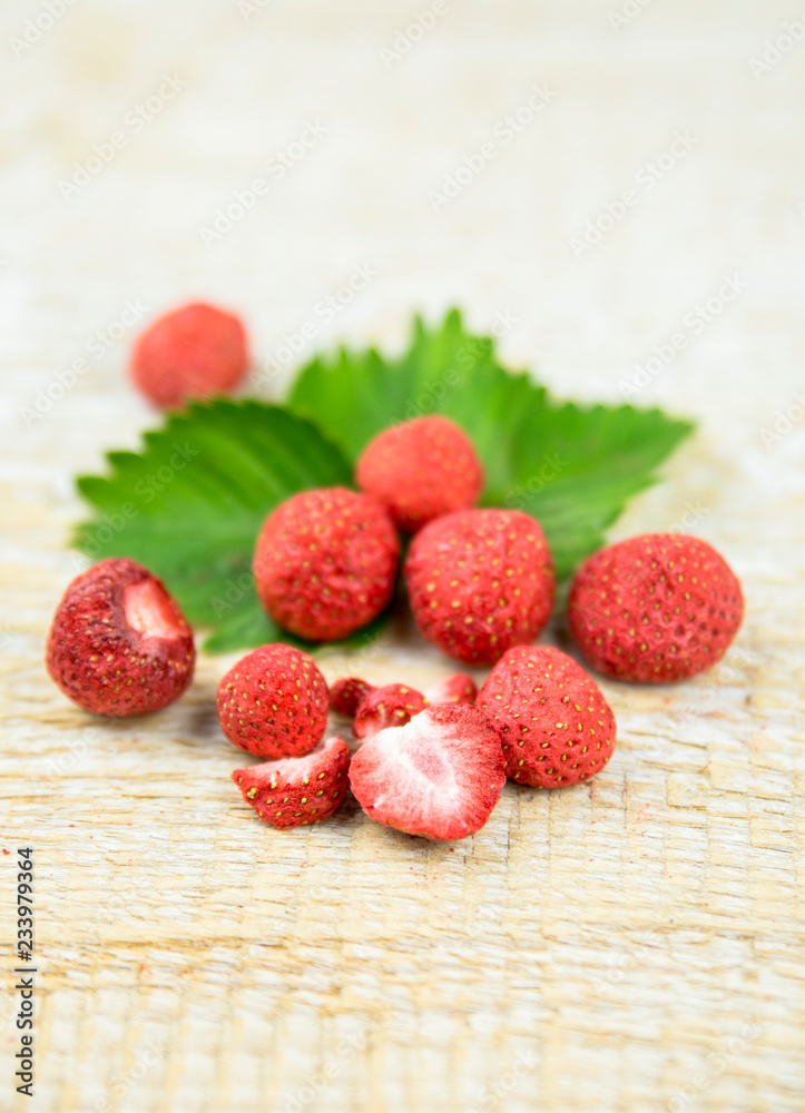 Freeze dried whole and pieces of strawberries on green strawberry leaf, healthy snack full of vitamins and nutrition on natural wooden background. Preserving berries for winter concept.