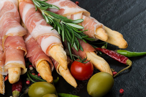 Grissini bread sticks with prosciutto on a black background with rosemary, pepper and olives