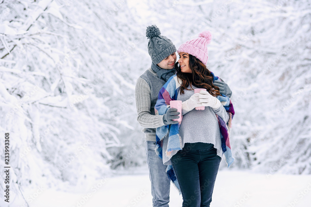 Beautiful pregnant woman with her husband dressed grey knitted sweaters, hat, scarf and jeans, holding pink cups with hot tea and walking in a snowy winter park on background.