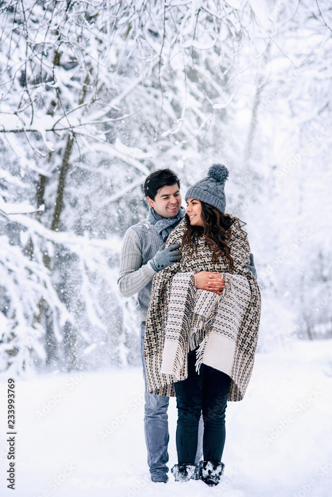 Young man hugs tenderly his beautiful pregnant wife covered in warm blanket with snowfall on background. Family love and care concept. Young man hugs tenderly his beautiful pregnant wife covered in warm blanket with snowfall on background. Family love and care concept.