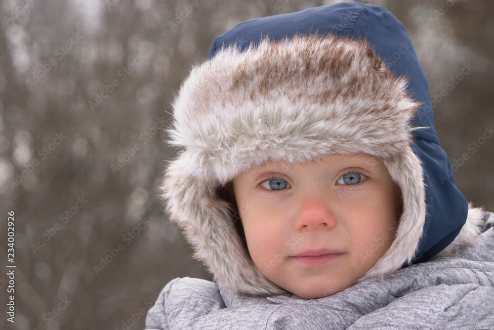 Cute little boy (toddler, two year old) in hat with fur trim and earflaps. Winter, cold weather, outdoor
