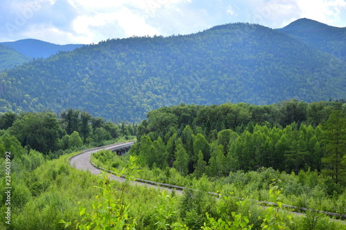 The road among green mountains. Asphalt road stretches among the emerald forest. In the background  dark blue mountains. Bright sunny day.