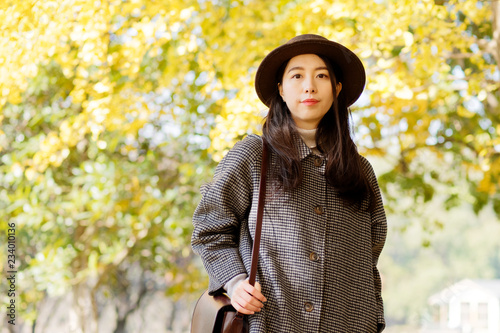 Portrait of young Chinese woman in fashionable coat and hat with golden autumn forest background in park, carring a bag. Cute girl in good mood posing in autumn day, enjoying good weather.