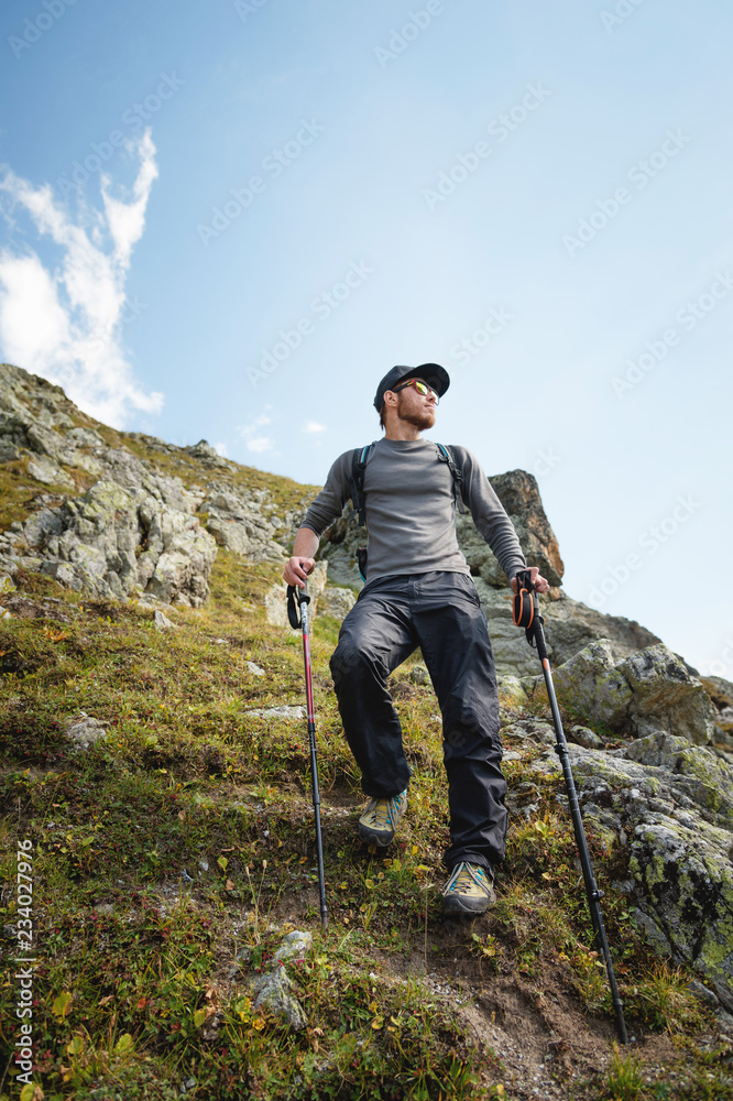 A bearded man in sunglasses and a cap with a backpack stands on top of a rock and looks into a rocky valley high in the mountains. The concept of tourism and easy trekking in the mountains outdoor