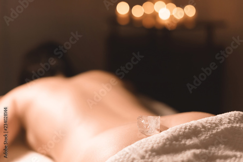 Close-up of a transparent ice cube lying on the lower back of a young girl lying on a couch for spa treatments against the backdrop of a dark room and burning candles. Spa treatments. Small DOF