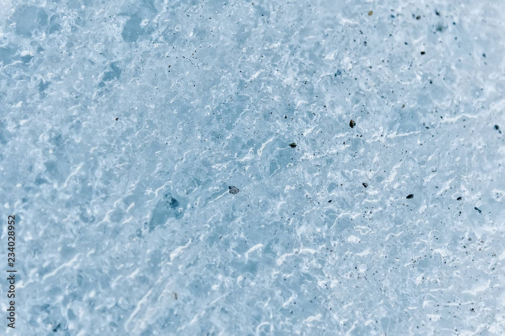 A close-up of the surface of a fragment of a glacier with a structure of strips and bubbles interspersed with sand and rocky rock. Ice blue texture to the light. Small DOF