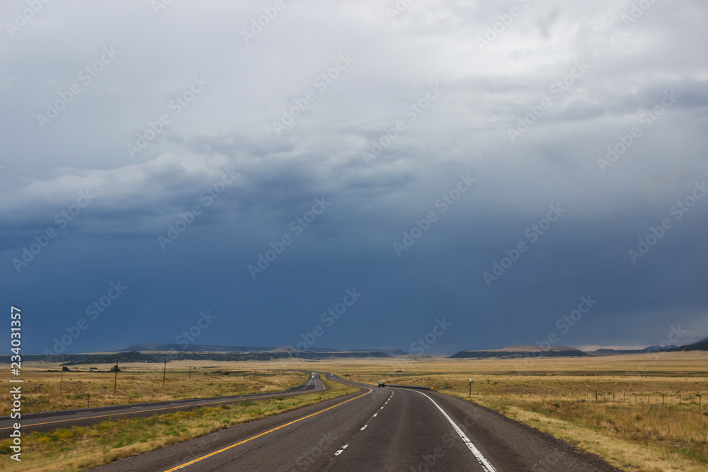 The desert highway among the steppe, in front of the blue mountains. Summer day, dark sky, big dark blue clouds in the sky