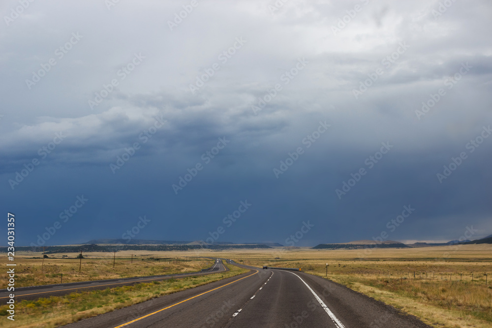 The desert highway among the steppe, in front of the blue mountains. Summer day, dark sky, big dark blue clouds in the sky The desert highway among the steppe, in front of the blue mountains. Summer day, dark sky, big dark blue clouds in the sky