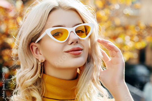 Outdoor close up portrait of young beautiful happy smiling blonde girl wearing white yellow cat eye sunglasses, hoop earrings, turtleneck, posing in street. Natural sunny day light