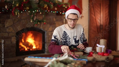 Bearded man sitting and tying a bow on a gifts for New Year near fireplace. Guy wearing Christmas hat wrapping present boxes in paper adding fir branches, cones, cane candies. Unmasculinity concept. photo