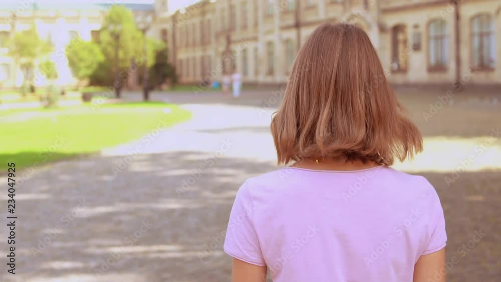 back view student walking in campus area turn to the camera with happy smile. girl with blond hair wearing casual t-shirt on the background beautiful old college back view student walking in campus area turn to the camera with happy smile. girl with blond hair wearing casual t-shirt on the background beautiful old college