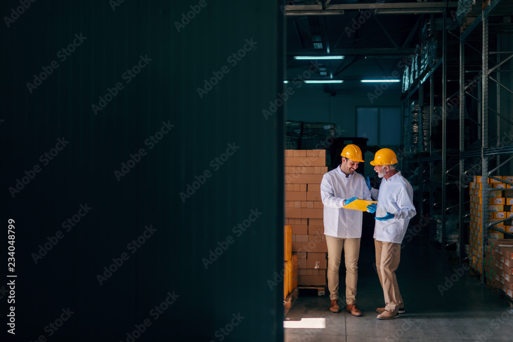 Two Caucasian workers discussing while younger worker holding documents. Warehouse interior.