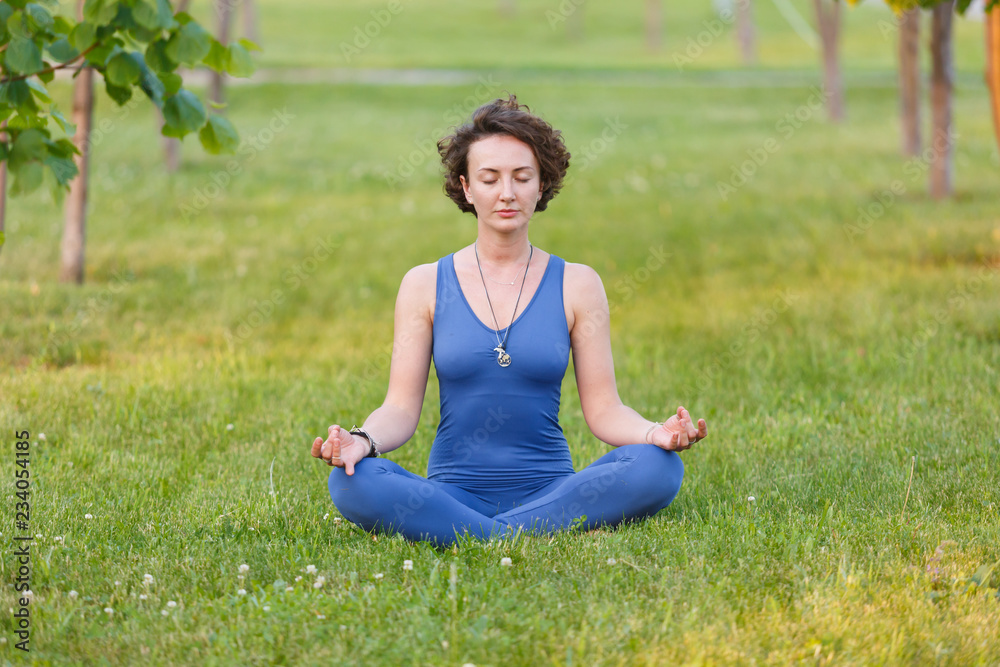Charming woman in gym suit meditating sitting in lotus pose with closed eyes on the lawn in the middle of the alley in the sunny summer day Charming woman in gym suit meditating sitting in lotus pose with closed eyes on the lawn in the middle of the alley in the sunny summer day