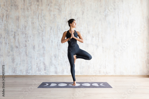Fototapeta Naklejka Na Ścianę i Meble -  Beautiful young brunette woman yoga instructor doing vrikshasana on a mat in a wooden floor standing in the gym with day lighting