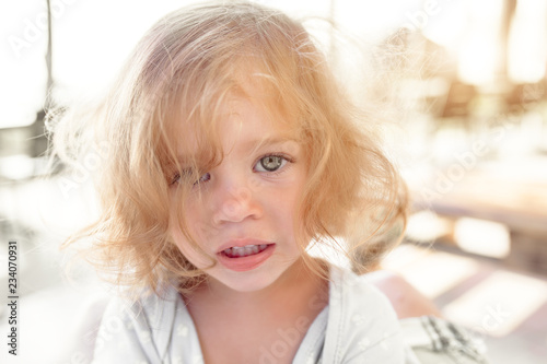 Close-up portrait of pretty young little blond pale unhappy moody friendless child girl looking sadly in camera on blurred sunny outdoors background. Children problems concept.