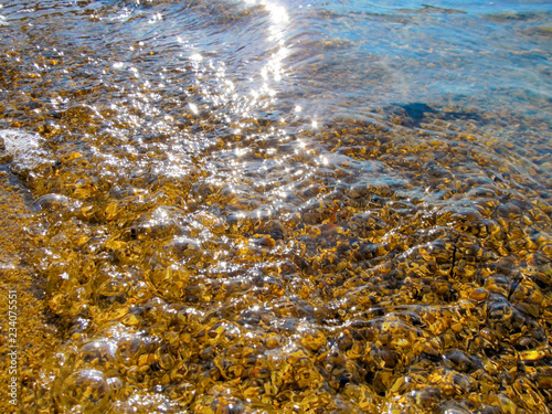Transparent water and pebbles beach - close-up. Summer background image of sunlight reflecting and glistening on the water surface and wet small stones in foreground.