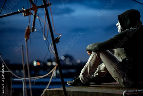 Mystery hoodie man in white mask sitting on rooftop of abandoned building looking up at the sky during the twilight time. Bipolar disorder or Major depressive disorder. Depression concept