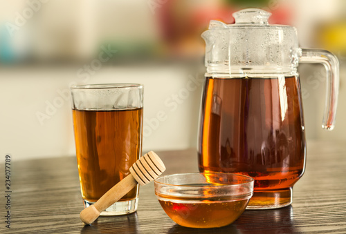 Pitcher with glass of hot black tea with bowl of honey on kitchen background.