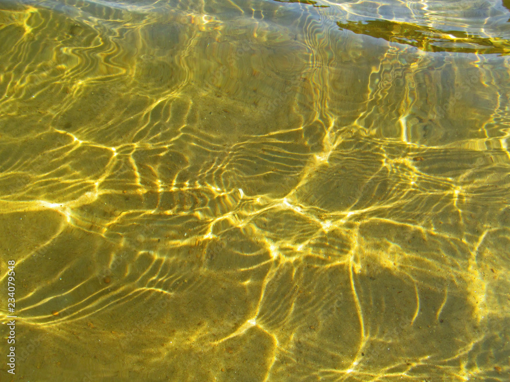 Transparent water surface with golden sun glare and sunny shining reflections on the sand. Top view of yellow ripple texture with sunlight refracting through liquid layer.