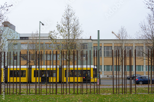 Fotografie Vertical steel pipes wall, part of Berlin wall memorial symbol divide the memorial park from street with background of Ubahn, yellow Tram and germany modern housing