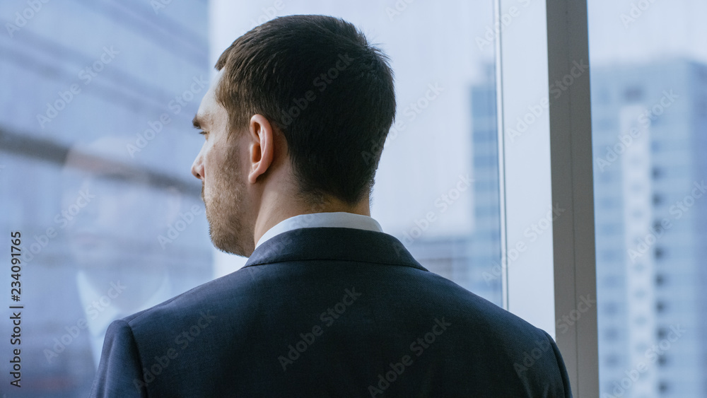 Close-up Shot of the Thoughtful Businessman in a Suit Standing in His Office and Contemplating Next Big Business Deal, Looking out of the Window. Big City Business District Panoramic Window View. Close-up Shot of the Thoughtful Businessman in a Suit Standing in His Office and Contemplating Next Big Business Deal, Looking out of the Window. Big City Business District Panoramic Window View.