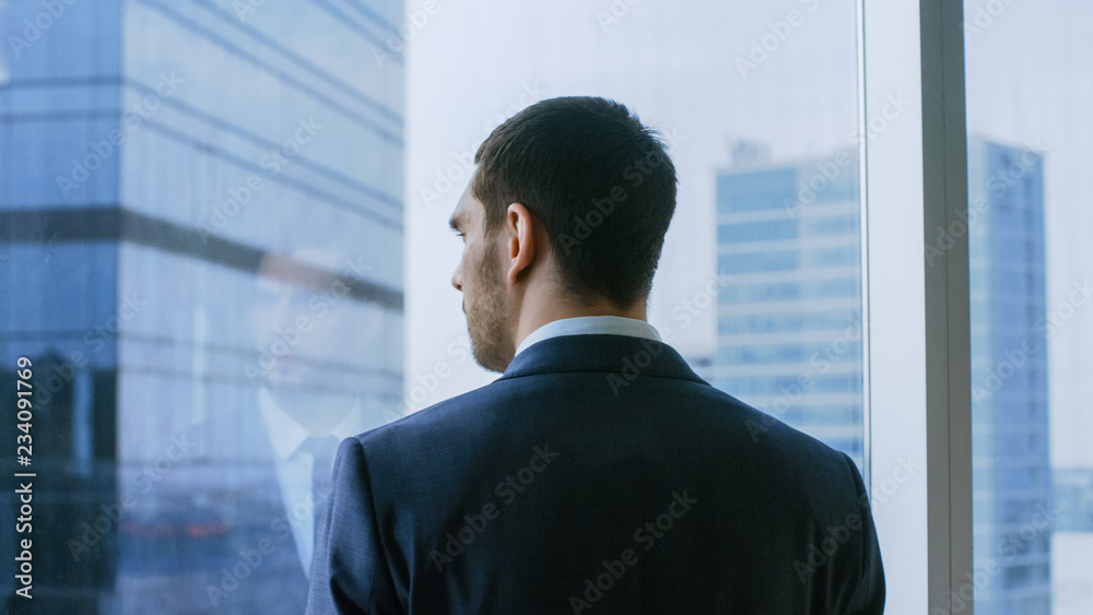 Medium Shot of the Thoughtful Businessman wearing a Suit Standing in His Office and Contemplating Next Big Business Deal, Looking out of the Window. Big City Business District Panoramic Window View. Medium Shot of the Thoughtful Businessman wearing a Suit Standing in His Office and Contemplating Next Big Business Deal, Looking out of the Window. Big City Business District Panoramic Window View.