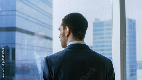 Medium Shot of the Thoughtful Businessman wearing a Suit Standing in His Office and Contemplating Next Big Business Deal, Looking out of the Window. Big City Business District Panoramic Window View.