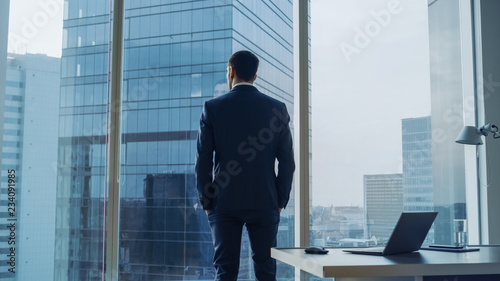 Back View of the Thoughtful Businessman wearing a Suit Standing in His Office, Hands in Pockets and Contemplating Next Big Business Deal, Looking out of the Window. Big City Panoramic Window View.
