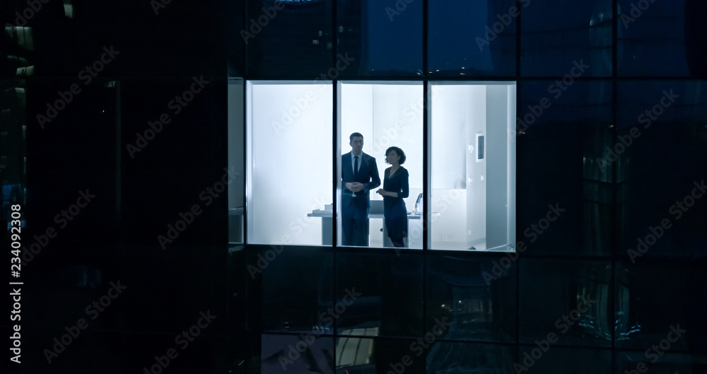 Aerial Shot From Outside of the Skyscraper Businessman and Businesswoman Talking while Looking Out of the Office Window. Shot of the Business District in the Evening and Businesspeople Working in City