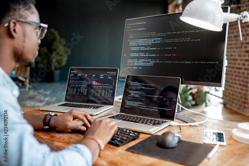 Young african male programmer writing program code sitting at the workplace with three monitors in the office. Image focused on the screen photo