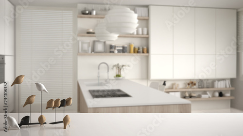 White table top or shelf with minimalistic bird ornament, birdie knick - knack over blurred white and wooden kitchen with cabinets, island with gas stove, modern interior design