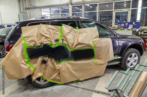 Partial painting of the side elements of the body of a black SUV car after an accident on the road, not damaged parts are covered with paper and tape from getting into paint in a car repair workshop