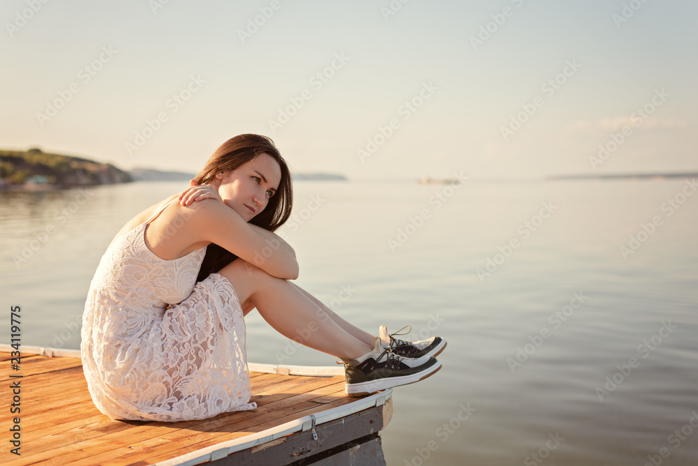 Sad girl sitting on the pier hugging her knees, looking into the distance, at sunset, loneliness, separation