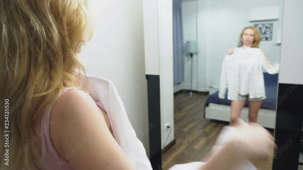 Clothing, wardrobe, fashion, style and concept of people. puzzled blonde makes a choice of clothes, standing near the closet and looking at herself in the mirror Clothing, wardrobe, fashion, style and concept of people. puzzled blonde makes a choice of clothes, standing near the closet and looking at herself in the mirror