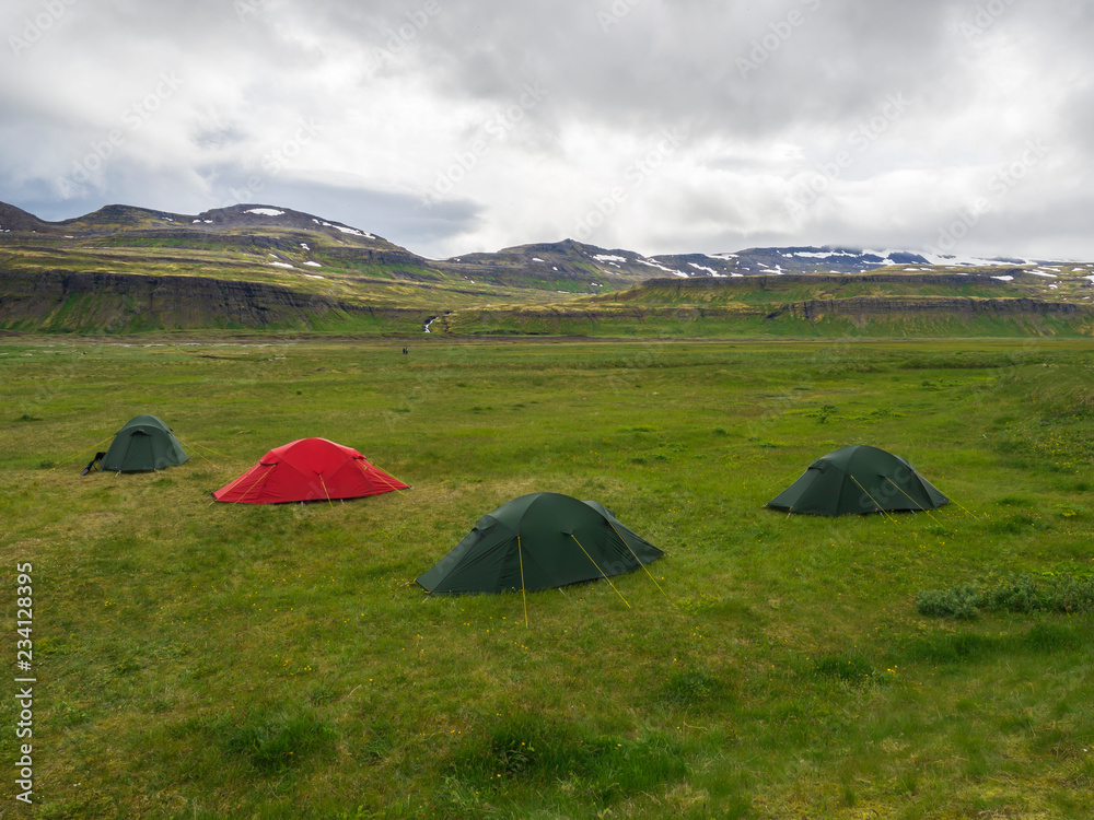 Hornvik campsite with three green tents and one red and view on beautiful Hornbjarg cliffs in remote nature reserve with green meadow, waterfall and hills, moody sky, Iceland, west fjords Hornstrandir